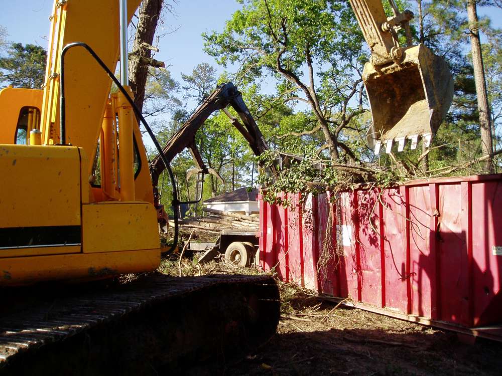 An excavator with a grapple loading cleared brush and tree limbs into a roll-off container for haul-off