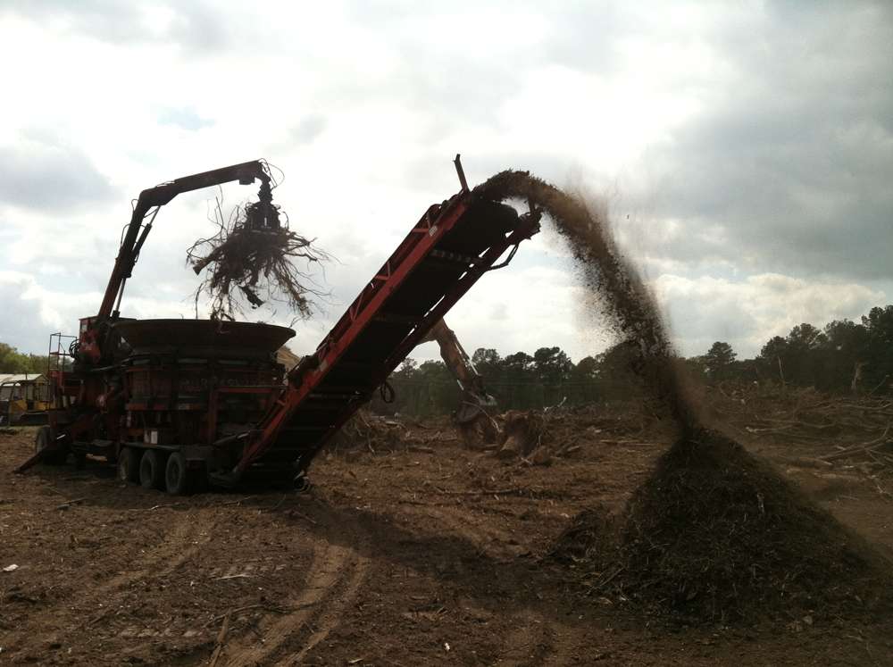 A horizontal grinder feeding cleared stumps and brush and blowing the ground material into a single mulch pile on a cleared site